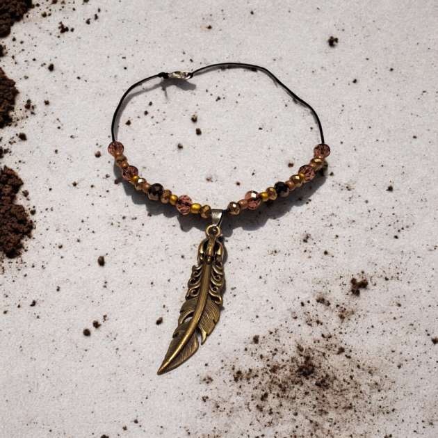 طوق يدوي بقلادة ريشة ذهبية وخرز كريستال بني موضوع على خلفية رملية